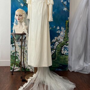 May include: Ivory vintage wedding dress with long sleeves and a high neckline. The dress features lace detailing on the bodice and cuffs, and a long, flowing train. A matching veil is displayed on a mannequin head. The background includes a floral painting.