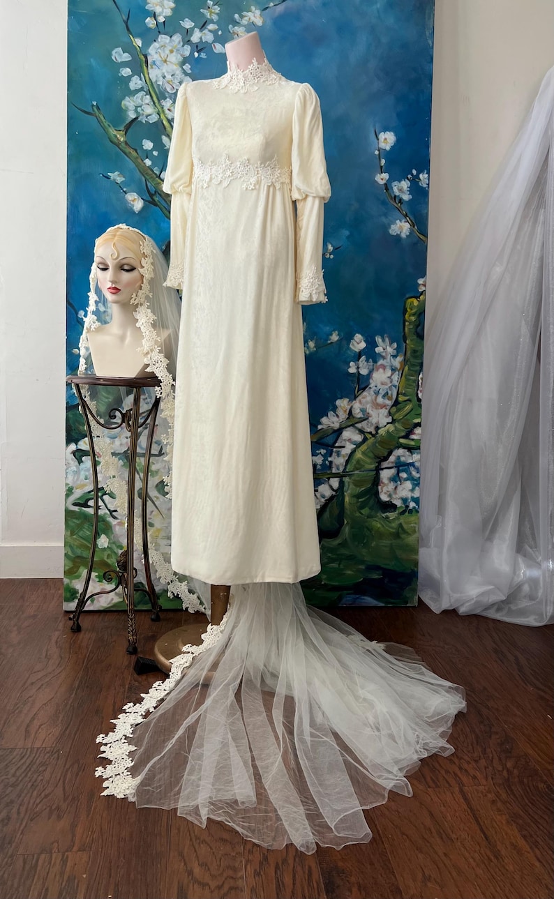 May include: Ivory vintage wedding dress with long sleeves and a high neckline. The dress features lace detailing on the bodice and cuffs, and a long, flowing train. A matching veil is displayed on a mannequin head. The background includes a floral painting.