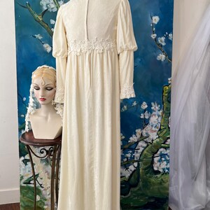 May include: Cream-colored vintage wedding dress with long sleeves and a high neckline. The dress features delicate lace detailing at the cuffs and waist. A mannequin head with a veil is on a small table to the left. The background is a blue painting with white flowers.