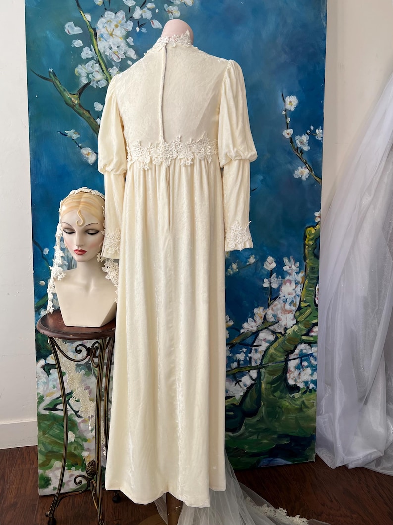 May include: Cream-colored vintage wedding dress with long sleeves and a high neckline. The dress features delicate lace detailing at the cuffs and waist. A mannequin head with a veil is on a small table to the left. The background is a blue painting with white flowers.