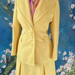 May include: A vintage yellow suit set on a mannequin. The suit features a blazer with three buttons and a pleated skirt. The suit has decorative stitching and is paired with a colorful scarf. The background is a painting of white flowers and blue sky.