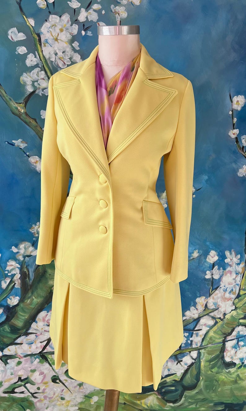 May include: A vintage yellow suit set on a mannequin. The suit features a blazer with three buttons and a pleated skirt. The suit has decorative stitching and is paired with a colorful scarf. The background is a painting of white flowers and blue sky.
