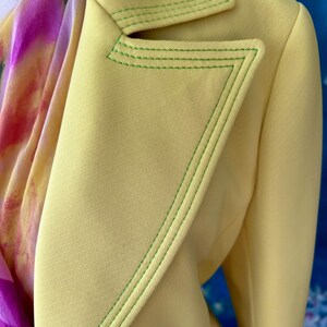 May include: A close-up of a light yellow blazer with green stitching along the lapels and edges. A colorful scarf with pink and purple hues is partially visible. The blazer has a tailored design.