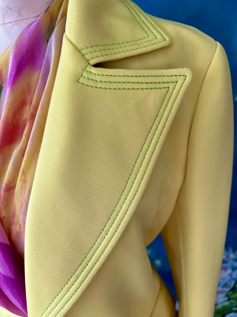 May include: A close-up of a light yellow blazer with green stitching along the lapels and edges. A colorful scarf with pink and purple hues is partially visible. The blazer has a tailored design.