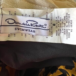 May include: White tag with black text that reads "Scandal Kenta Swimwear" and size information. The tag is attached to a black and brown fabric.