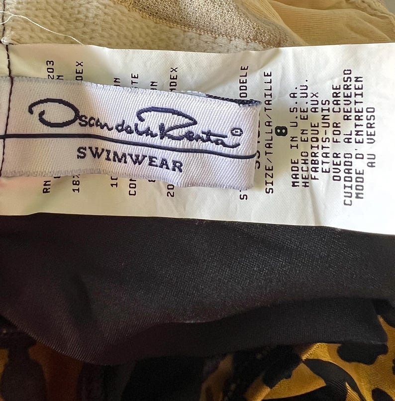 May include: White tag with black text that reads "Scandal Kenta Swimwear" and size information. The tag is attached to a black and brown fabric.