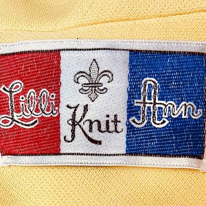 May include: A rectangular label with the words "Lilli Knit Ann" embroidered in white script on a yellow background. The label is divided into three vertical sections: red, white, and blue. A fleur-de-lis is in the white section.