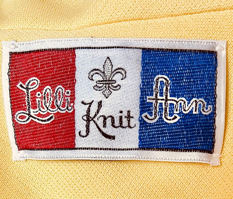 May include: A rectangular label with the words "Lilli Knit Ann" embroidered in white script on a yellow background. The label is divided into three vertical sections: red, white, and blue. A fleur-de-lis is in the white section.