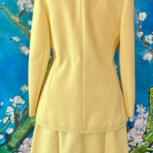 May include: A pale yellow two-piece suit, consisting of a long-sleeved blazer and a knee-length skirt. The blazer has a tailored fit with a subtle seam detail and a matching skirt with a pleated design. The suit is made of a smooth fabric.