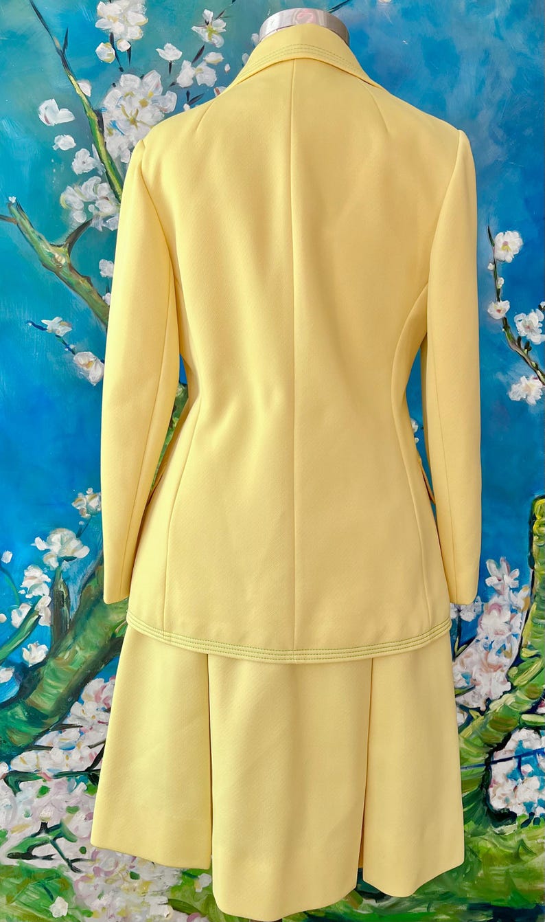 May include: A pale yellow two-piece suit, consisting of a long-sleeved blazer and a knee-length skirt. The blazer has a tailored fit with a subtle seam detail and a matching skirt with a pleated design. The suit is made of a smooth fabric.