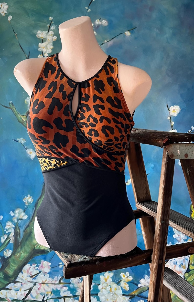 May include: A black and orange leopard print swimsuit with a keyhole cutout at the neckline. The swimsuit has a black bottom and a thin orange band at the waist.