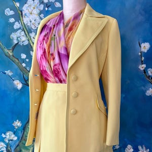 May include: A pale yellow skirt suit with a matching double-breasted jacket. The jacket has three buttons and a decorative stitch. A colorful scarf with pink and purple floral patterns is draped around the neck. The suit is displayed on a mannequin.