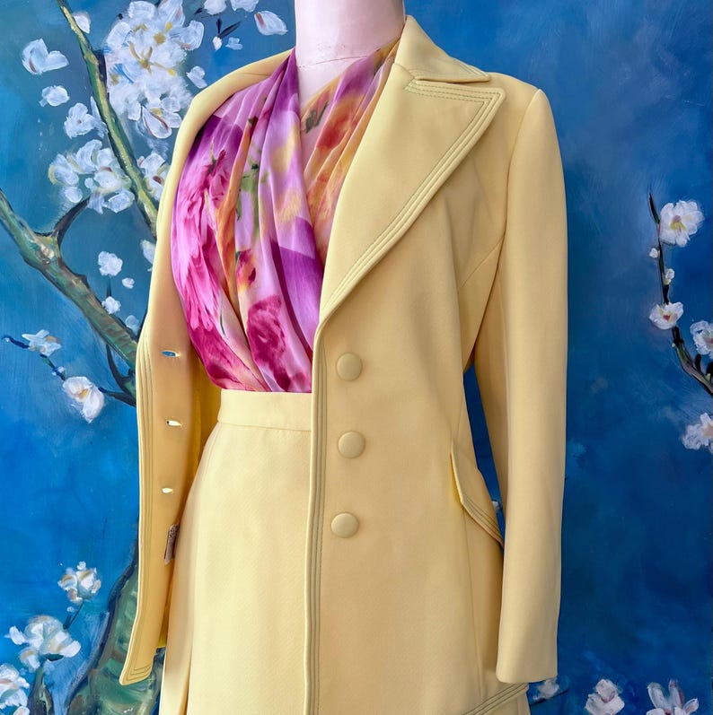 May include: A pale yellow skirt suit with a matching double-breasted jacket. The jacket has three buttons and a decorative stitch. A colorful scarf with pink and purple floral patterns is draped around the neck. The suit is displayed on a mannequin.
