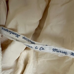 May include: Close-up of a cream-colored fabric with a white ribbon featuring the text "Murray Hamburger" and "nger & Co." in navy blue. The fabric appears to be part of a garment.