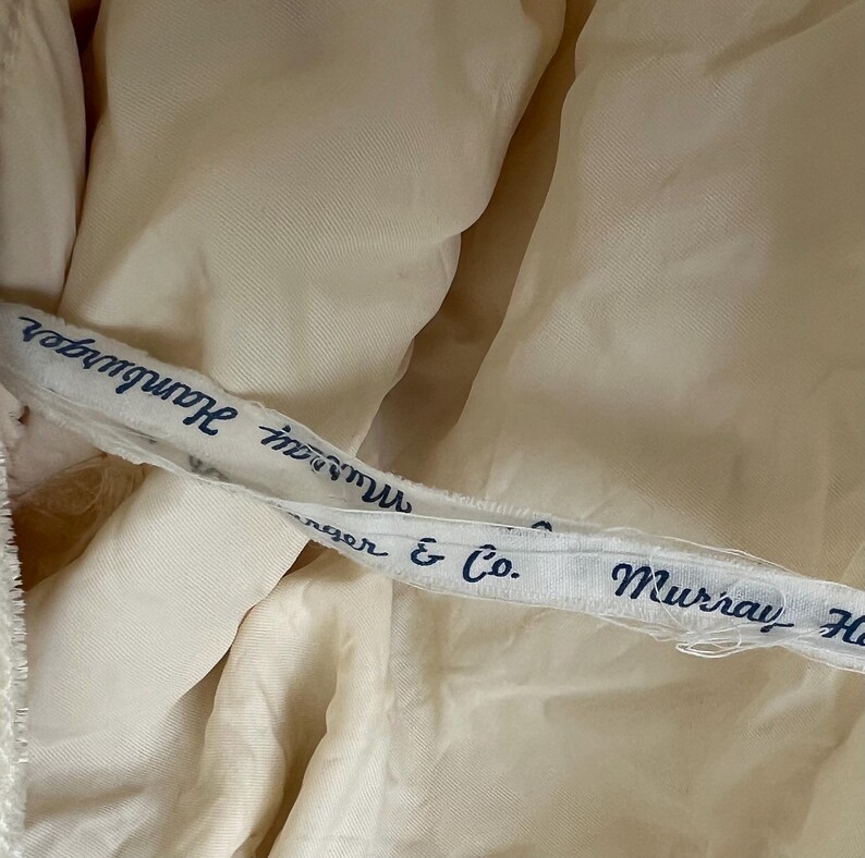 May include: Close-up of a cream-colored fabric with a white ribbon featuring the text "Murray Hamburger" and "nger & Co." in navy blue. The fabric appears to be part of a garment.