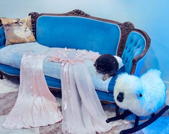May include: A light pink satin slip dress and a matching sheer pink robe with long sleeves and a tie-front closure. The garments are displayed on a blue velvet sofa alongside a small black dog and a white plush rocking sheep toy.