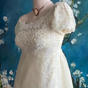 May include: An off-white dress featuring short puff sleeves, a scoop neckline, and an empire waist. The bodice and sleeves are adorned with white floral lace appliqués and scattered small white beads. A cameo necklace is worn around the neck.