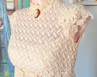 May include: A cream-colored sleeveless dress featuring an intricate leaf-patterned embroidery throughout. The garment has a high lace neckline adorned with a small oval cameo brooch and floral lace detailing at the shoulders and waist.