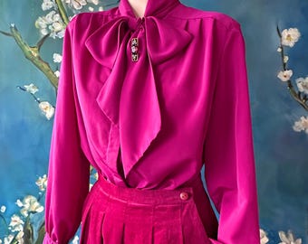 May include: A vibrant fuchsia blouse with a large bow and a matching pleated skirt. The long-sleeved blouse features a high collar and a decorative brooch. The skirt has a button closure. The outfit is displayed on a mannequin.