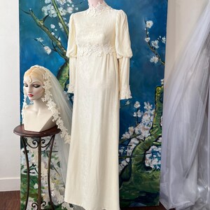 May include: Ivory-colored vintage wedding dress with long sleeves and lace detailing. The dress is displayed on a mannequin, with a matching veil on a bust nearby. The dress has a high neckline and a flowing skirt, perfect for a classic bridal look.