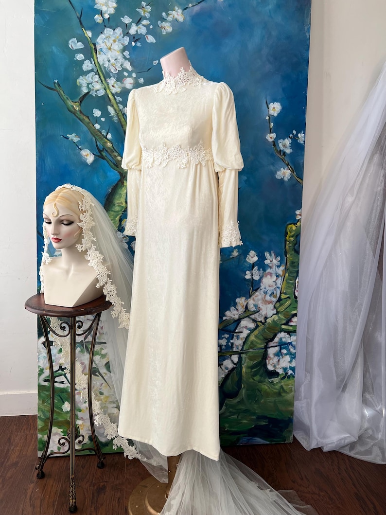 May include: Ivory-colored vintage wedding dress with long sleeves and lace detailing. The dress is displayed on a mannequin, with a matching veil on a bust nearby. The dress has a high neckline and a flowing skirt, perfect for a classic bridal look.
