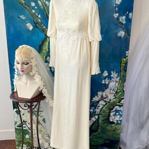 May include: Ivory vintage wedding dress with long sleeves, a high neckline, and lace detailing at the cuffs, waist, and neckline. A mannequin head with a veil is on a small table. The background features a blue and white floral painting.