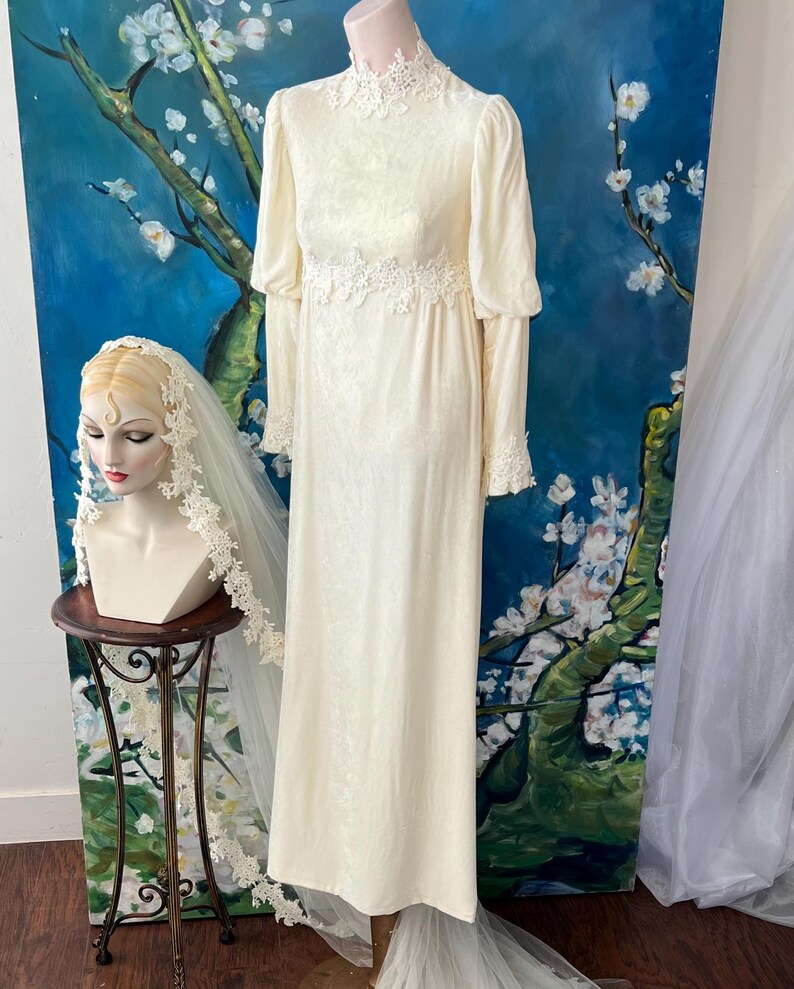May include: Ivory vintage wedding dress with long sleeves, a high neckline, and lace detailing at the cuffs, waist, and neckline. A mannequin head with a veil is on a small table. The background features a blue and white floral painting.