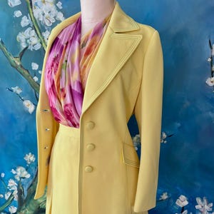 May include: A pale yellow suit with a matching skirt and jacket. The jacket has a double-breasted design with three buttons and a notched collar. A colorful scarf with pink and yellow floral patterns is draped around the neck. The suit is displayed on a mannequin.