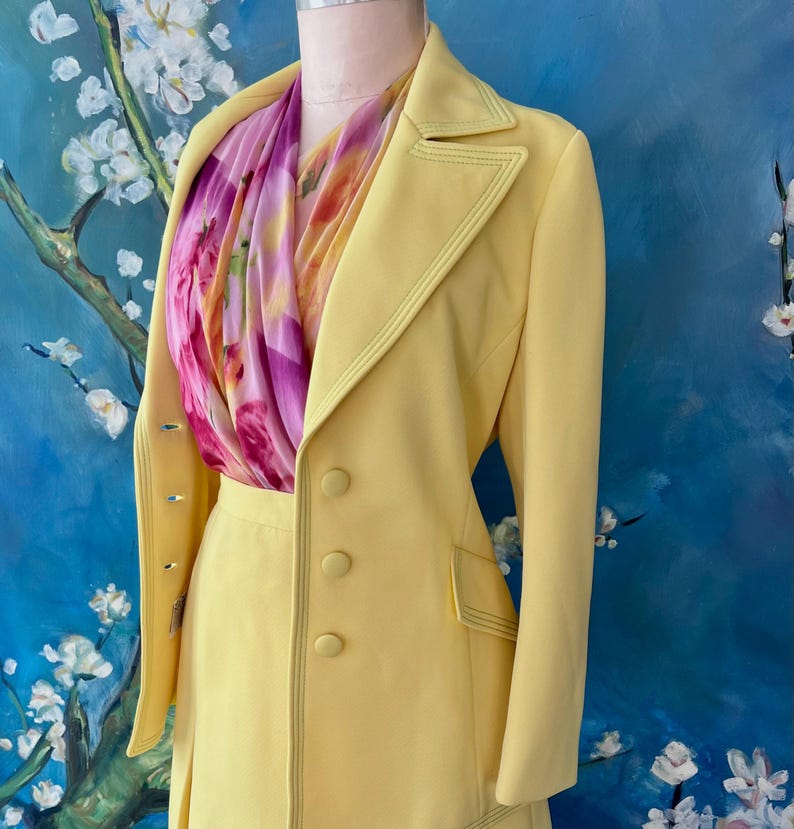 May include: A pale yellow suit with a matching skirt and jacket. The jacket has a double-breasted design with three buttons and a notched collar. A colorful scarf with pink and yellow floral patterns is draped around the neck. The suit is displayed on a mannequin.