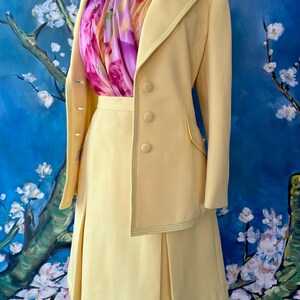 May include: A pale yellow skirt suit with a matching blazer and pleated skirt. The blazer has three buttons and a decorative stitch. A colorful scarf with pink and purple floral patterns is worn underneath the blazer.