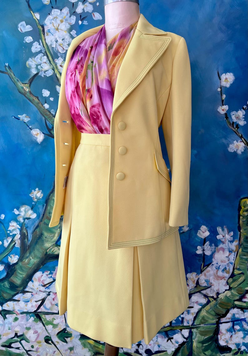 May include: A pale yellow skirt suit with a matching blazer and pleated skirt. The blazer has three buttons and a decorative stitch. A colorful scarf with pink and purple floral patterns is worn underneath the blazer.