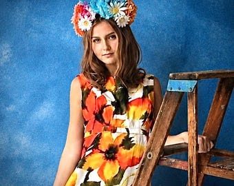 May include: A floral crown featuring a mix of orange, blue, and white daisies and other blossoms worn on the head. The accessory is paired with a sleeveless dress showcasing a vibrant, large-scale orange, yellow, and white floral print pattern.