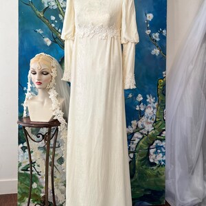 May include: Ivory vintage wedding dress with long sleeves and a high neckline. The dress features delicate lace detailing at the cuffs, neckline, and waist. A mannequin head with a veil is on the left. The dress is displayed in front of a blue and white floral painting.
