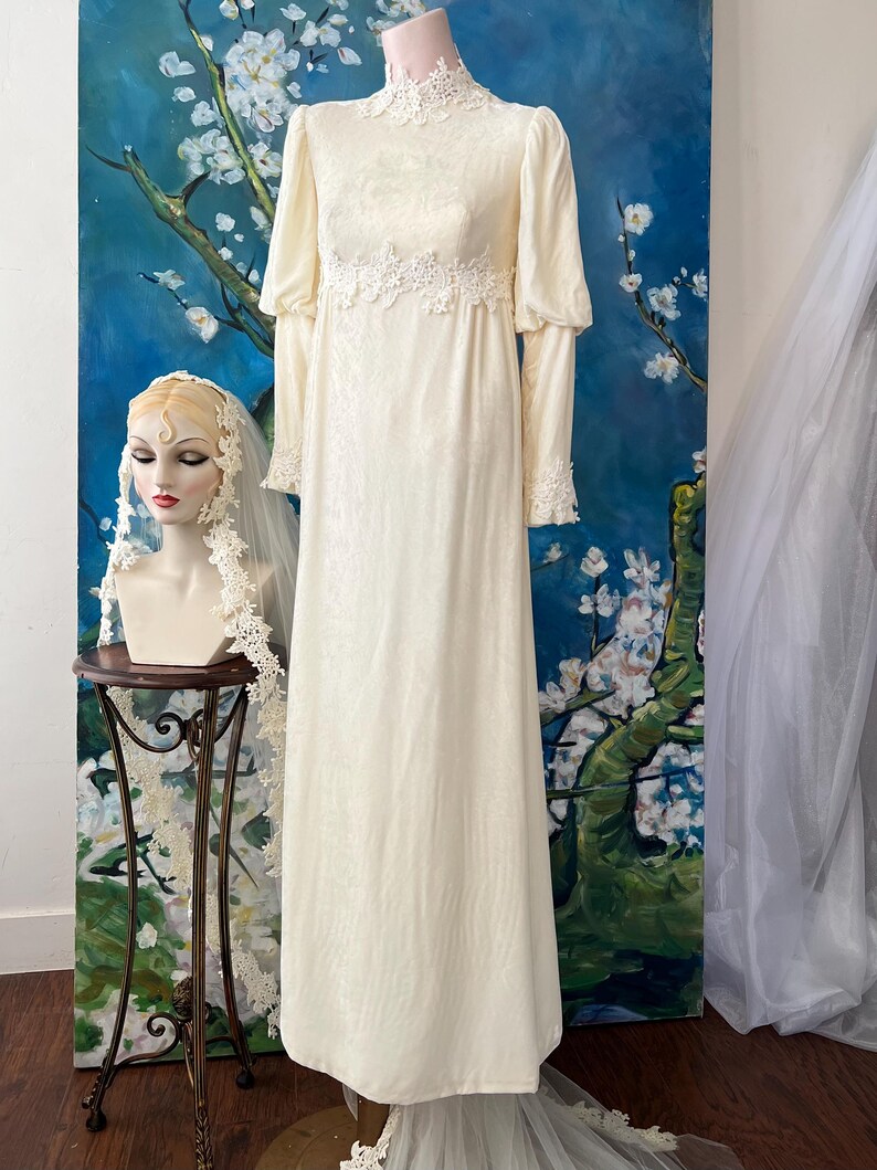 May include: Ivory vintage wedding dress with long sleeves and a high neckline. The dress features delicate lace detailing at the cuffs, neckline, and waist. A mannequin head with a veil is on the left. The dress is displayed in front of a blue and white floral painting.