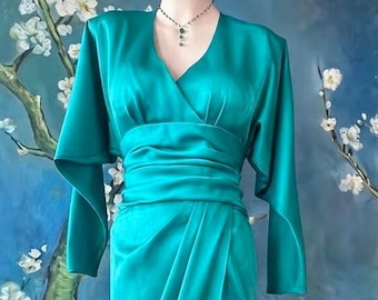 May include: A vibrant teal satin dress featuring a V-neckline, long sleeves with a draped silhouette, and a wide, gathered waistband. The garment is paired with a delicate necklace featuring small dark beads on a thin chain.