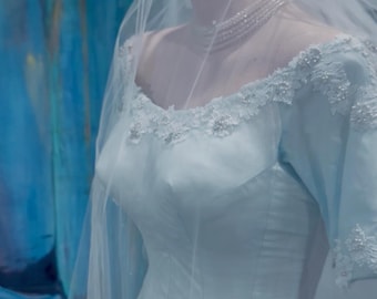 May include: A light blue wedding dress on a mannequin, featuring a sweetheart neckline and delicate lace detailing. The dress is adorned with sparkling embellishments and a sheer veil. A silver tiara and layered necklace complete the bridal look.