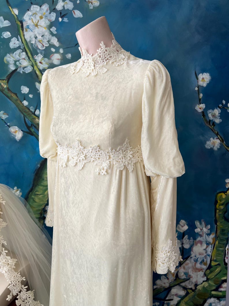 May include: Ivory-colored vintage wedding dress with long sleeves and a high neckline. The dress features delicate lace trim at the neckline, waist, and cuffs. The dress is displayed on a mannequin against a blue background with a floral painting.