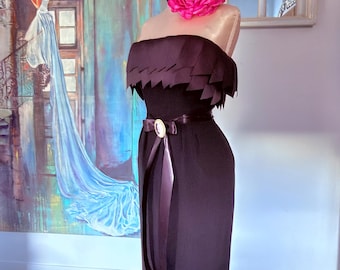 May include: A strapless black dress featuring a layered, jagged-edge bodice and a matching hemline. The dress is cinched at the waist with a dark satin ribbon belt that includes a central oval brooch. A large, vibrant hot pink fabric flower is pinned.