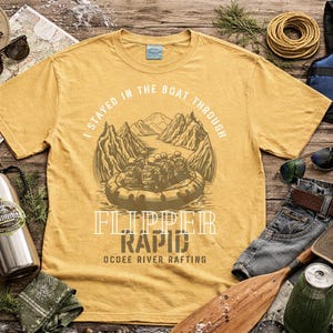 Può includere: T-shirt giallo senape con grafica di rafting e la scritta "I stayed in the boat through Flipper Rapid Ocoee River Rafting." Accessori: cappello, occhiali da sole, borraccia e giubbotto di salvataggio.