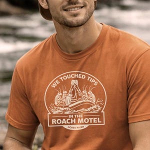 Può includere: T-shirt arancione a maniche corte con grafica bianca che recita "We Touched Tips - In The Roach Motel - Ocoee River." La persona nell'immagine indossa un cappellino da camionista e tiene in mano una borraccia verde.
