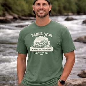Può includere: T-shirt verde con la scritta "TABLE SAW" sopra un disegno di una lama di sega. Sotto il disegno, il testo recita "NOT OSHA COMPLIANT OCOEE RIVER". La persona indossa un berretto verde e un orologio.