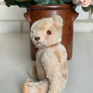 Op de afbeelding: Een vintage, crèmekleurige teddybeer met een rode strik zit op een wit oppervlak. De beer heeft een knoopneus en zwarte ogen.