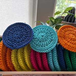 May include: A wooden box filled with a variety of colorful, round, crocheted items. The colors include green, orange, yellow, pink, blue, and teal. The items are arranged in a neat stack, showcasing the handmade texture.
