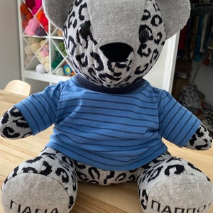 May include: A stuffed bear with a grey and black leopard print pattern on its head, arms, and legs. It wears a blue t-shirt with thin horizontal black stripes. The bear has black eyes and nose, and the text "ΓΙΑΓΙΑ" and "ΠΑΠΠΟΥ" on its feet.