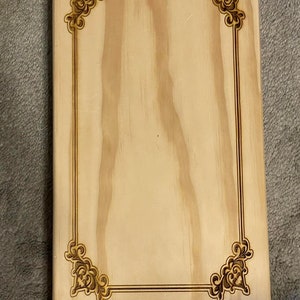 May include: A rectangular wooden plaque with a light wood grain finish. The plaque features a decorative border and corner accents, all in a darker, burnt wood color. The design is symmetrical and ornate.