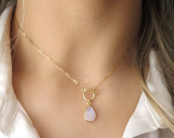 May include: A gold-colored necklace with a teardrop-shaped pendant. The pendant has a light blue and white stone. The necklace is fastened with a toggle clasp and is worn against a white shirt.