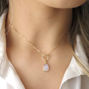 May include: A gold-colored necklace with a teardrop-shaped pendant. The pendant has a light blue and white stone. The necklace is fastened with a toggle clasp and is worn against a white shirt.