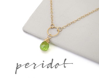 May include: A delicate gold chain necklace with a small green peridot gemstone pendant. The pendant is wrapped in gold wire and hangs from a gold circle.