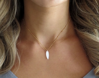 May include: A gold chain necklace with a white, teardrop-shaped pendant. The necklace is worn by a person with long, blonde hair.