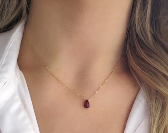 May include: A delicate gold chain necklace with a small, teardrop-shaped red gemstone pendant.